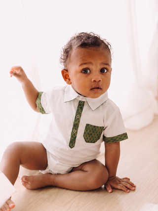 A baby sitting on a light wood floor wearing a white short-sleeve collared romper with green patterned trim on the collar placket, sleeve cuffs, and chest pocket.