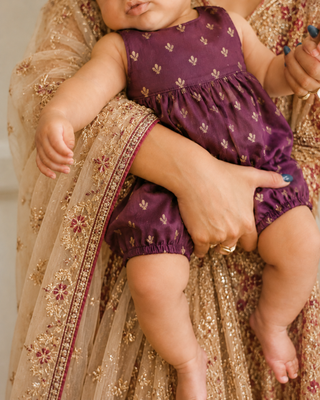 A baby wearing a purple sleeveless romper with small gold floral motifs, held by an adult in a beige and gold embroidered traditional outfit.