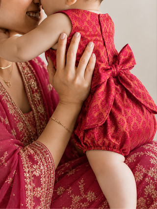 A close-up lifestyle shot of a baby wearing a bright red patterned romper with a large bow on the back, held by a mother wearing a matching red and gold embroidered sari.