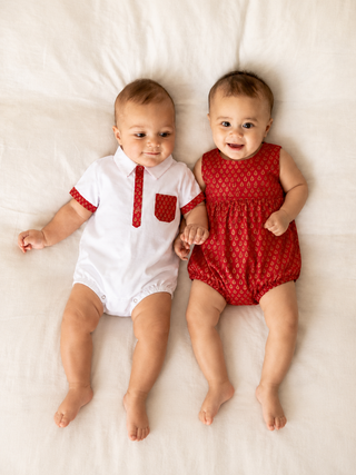 Two babies lying side by side on a white bed, wearing matching red and white patterned baby clothes. The baby on the left wears a white short-sleeve bodysuit with red trim and a red tie detail, and the baby on the right wears a red patterned sleeveless romper.