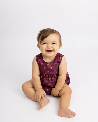 Baby sitting on a white background wearing a purple romper with a floral pattern.