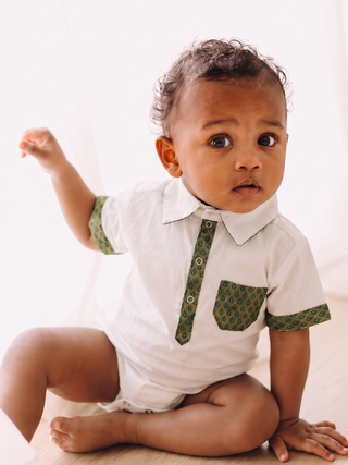 A baby sitting on a light wood floor wearing a white short-sleeve collared romper with green patterned trim on the collar placket, sleeve cuffs, and chest pocket.