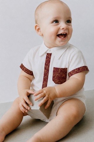 A smiling baby sitting on a light gray floor wearing a white short-sleeve collared bodysuit with a burgundy patterned front placket and pocket, holding a gray block.