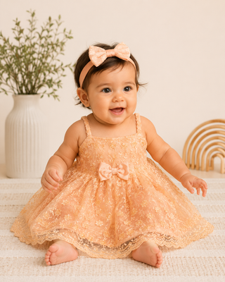 Baby in a peach lace dress with a bow sitting on a soft surface.