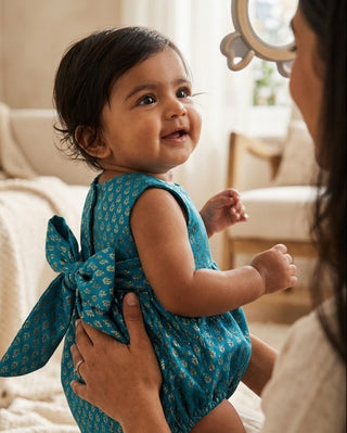 A baby wearing a teal blue patterned romper with a large matching bow on the back, held by an adult in a softly lit bedroom setting.