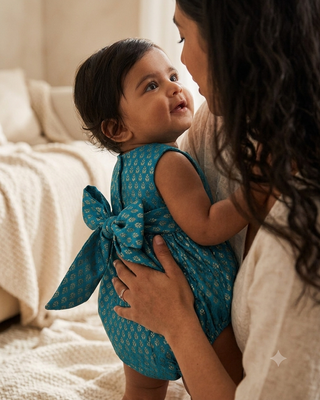 A toddler wearing a teal blue patterned romper with a large bow on the back stands on a bed while being held by an adult. The background shows a cozy, softly lit bedroom setting with neutral-colored bedding.