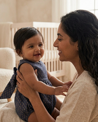 A smiling baby in a navy patterned romper sits on a woman's lap in a softly lit nursery. The woman wears a light beige linen top, and a cream-colored crib and armchair are visible in the background.